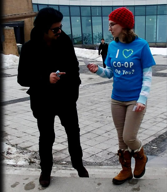 A student wearing an "I Love Co-Op" shirt passes out stickers in front of the Student Life Centre.