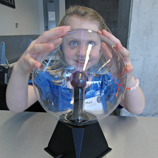 A girl smiles as she places her hands on a plasma ball and lightning plays inside.